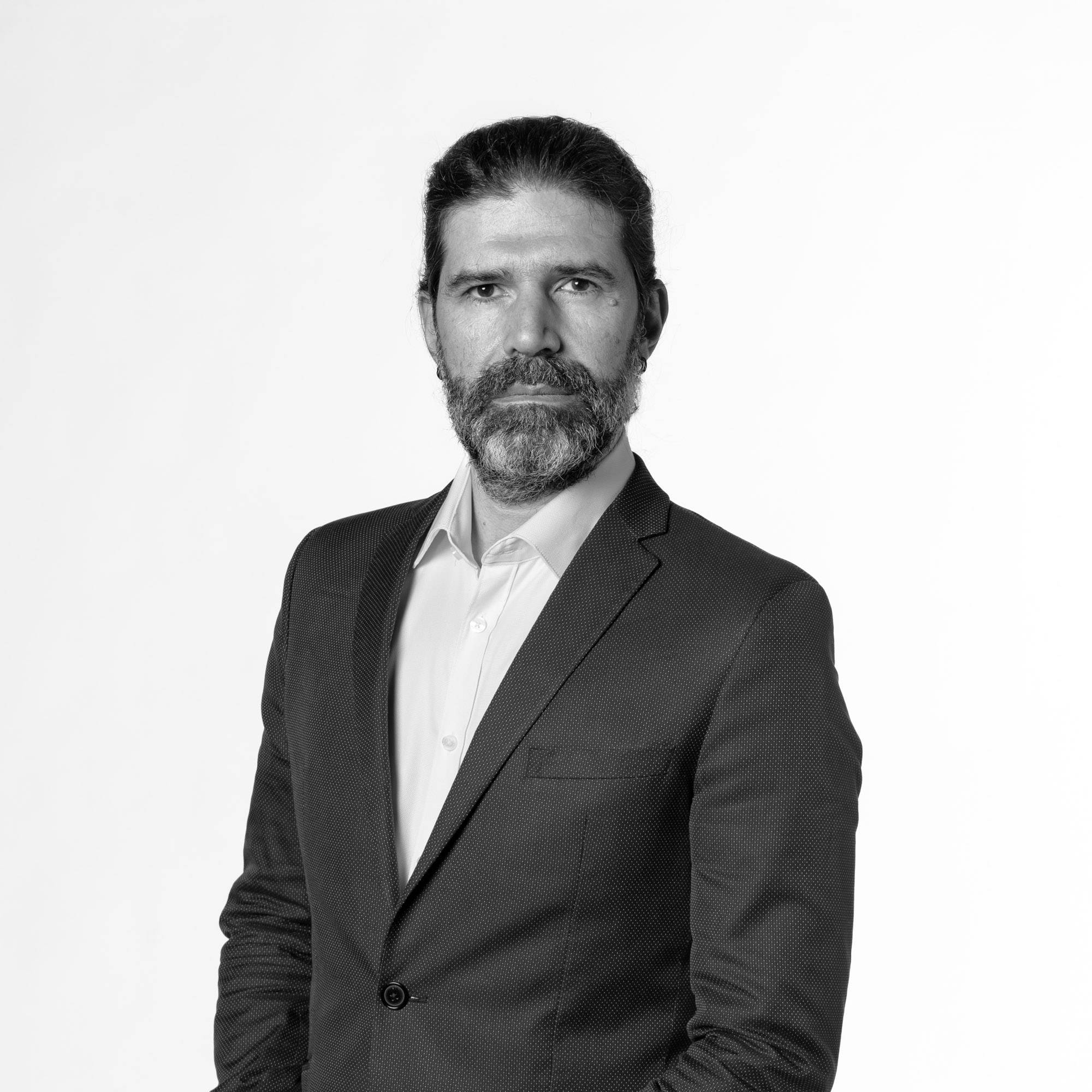 Un hombre con barba y cabello largo viste un traje oscuro y se presenta de manera seria frente a un fondo blanco.