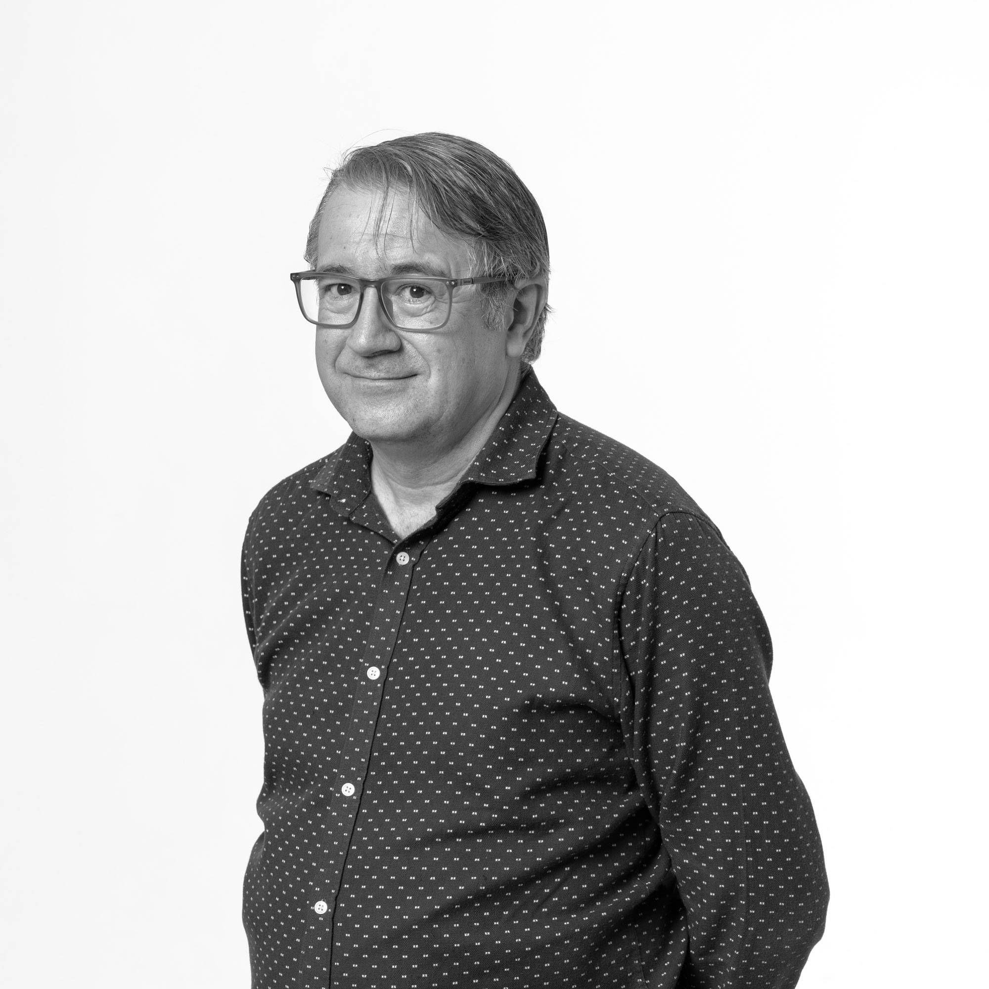 Retrato de un hombre de pie, con gafas y camisa con puntos sobre un fondo blanco.