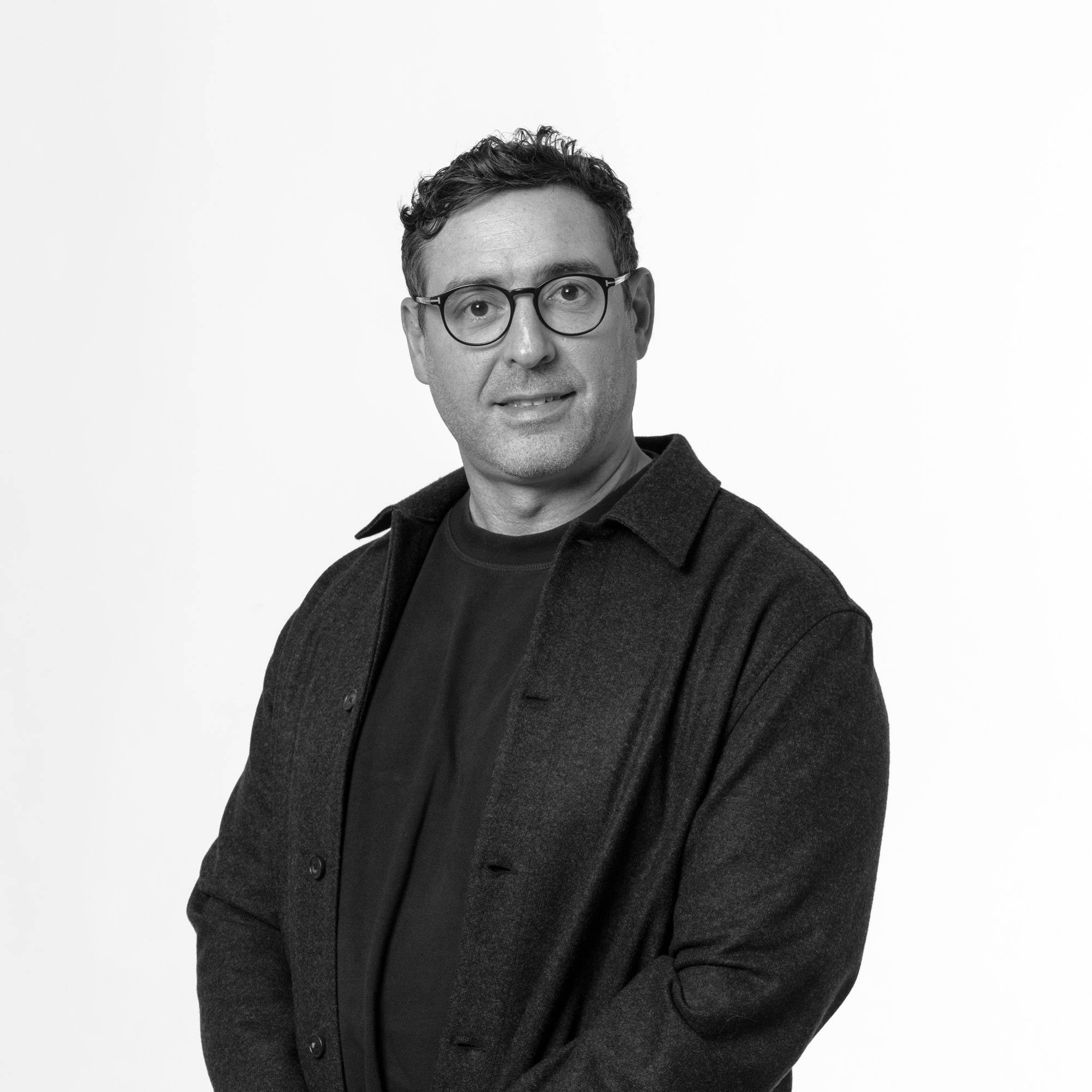 Un hombre de pie con gafas y una camisa negra en un fondo blanco.