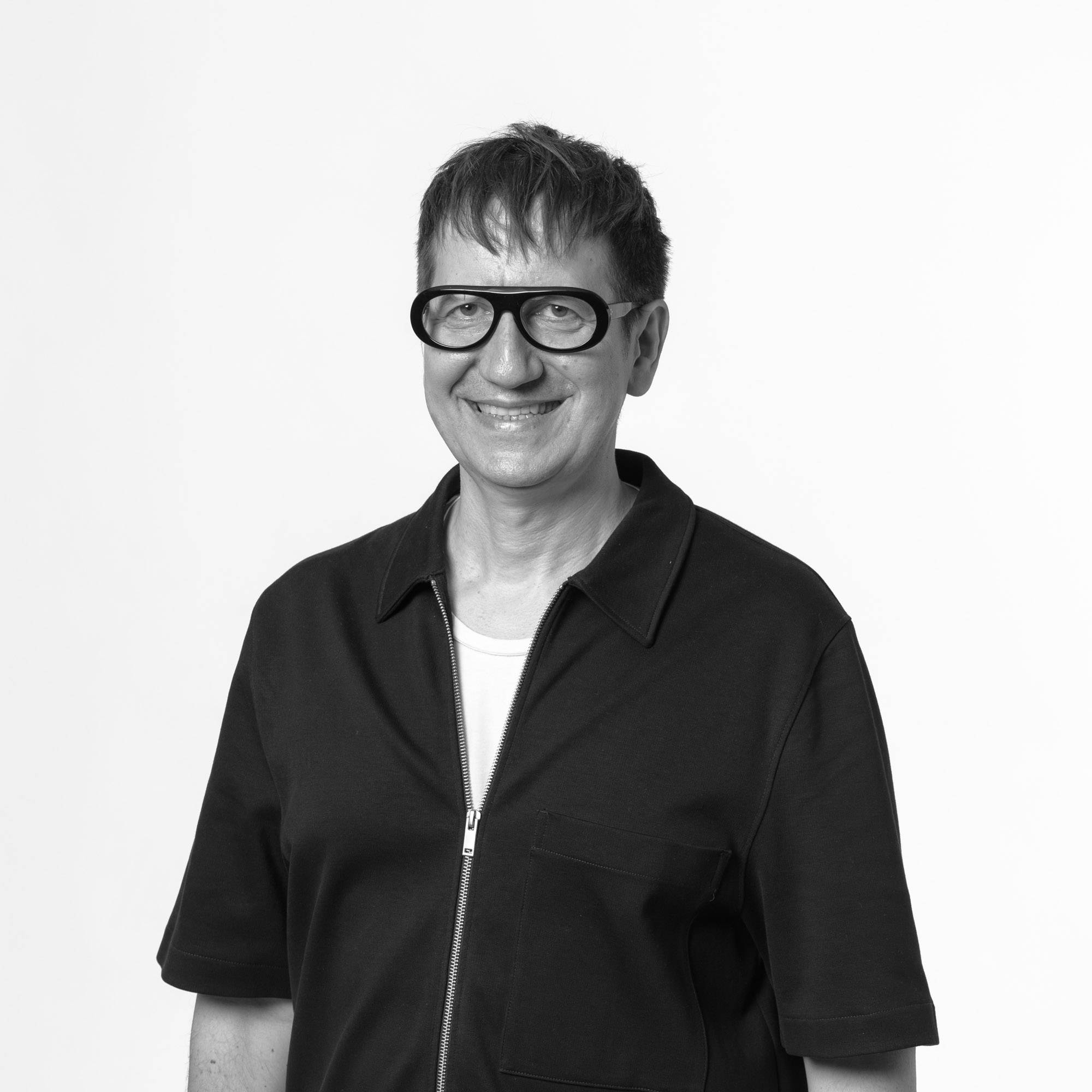 Una persona sonriente de pie, vestida con una camiseta blanca y una chaqueta oscura, frente a un fondo blanco.