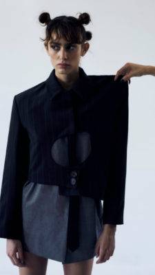 Una modelo viste una chaqueta negra en combinación con una prenda gris, mientras alguien sostiene la chaqueta.