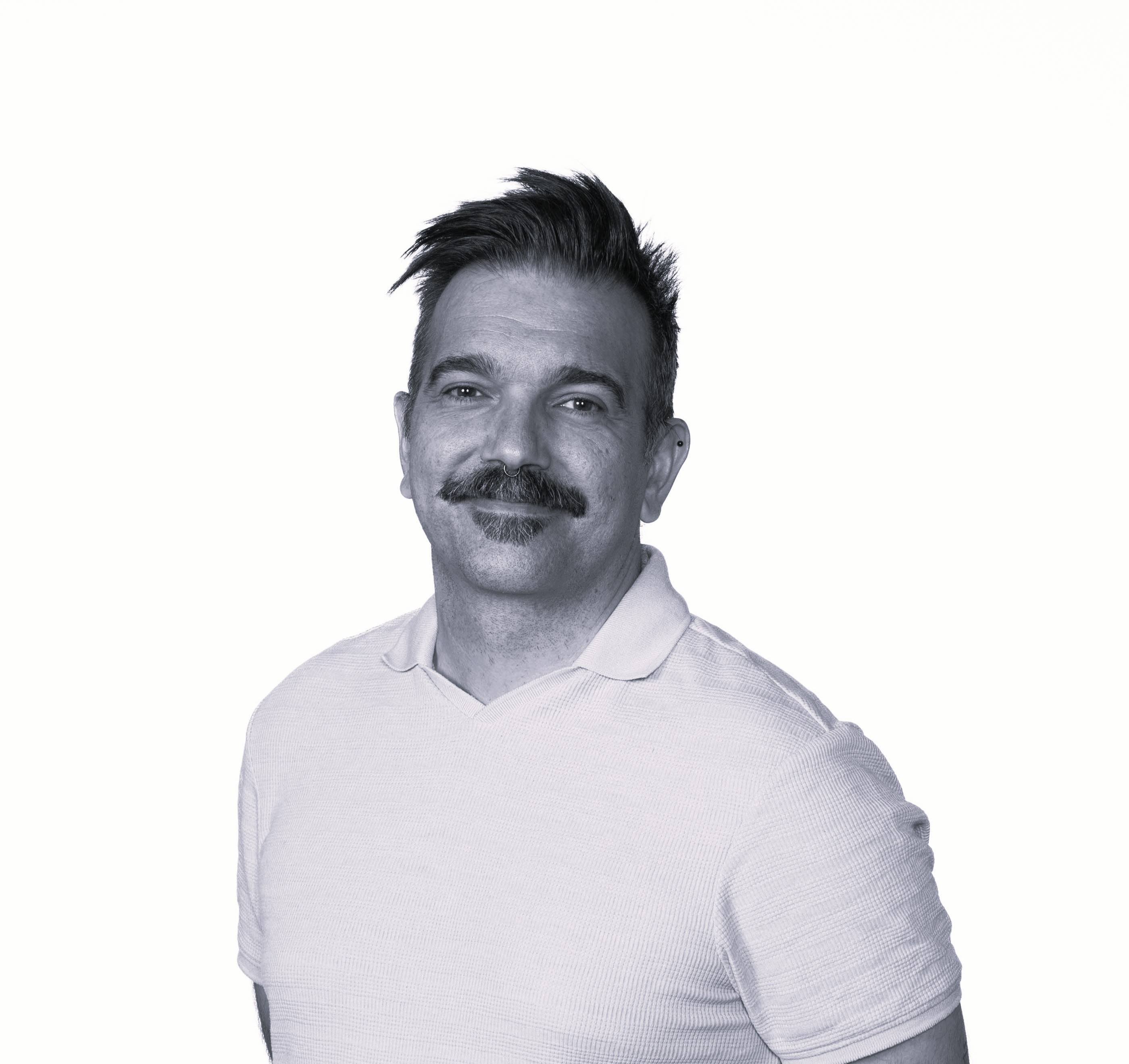 Un hombre con bigote y cabello corto, vestido con una camiseta de color claro, sonríe frente a un fondo blanco.