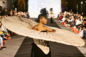 Una modelo presenta un vestido de gran tamaño en una pasarela con público sentado a los lados.