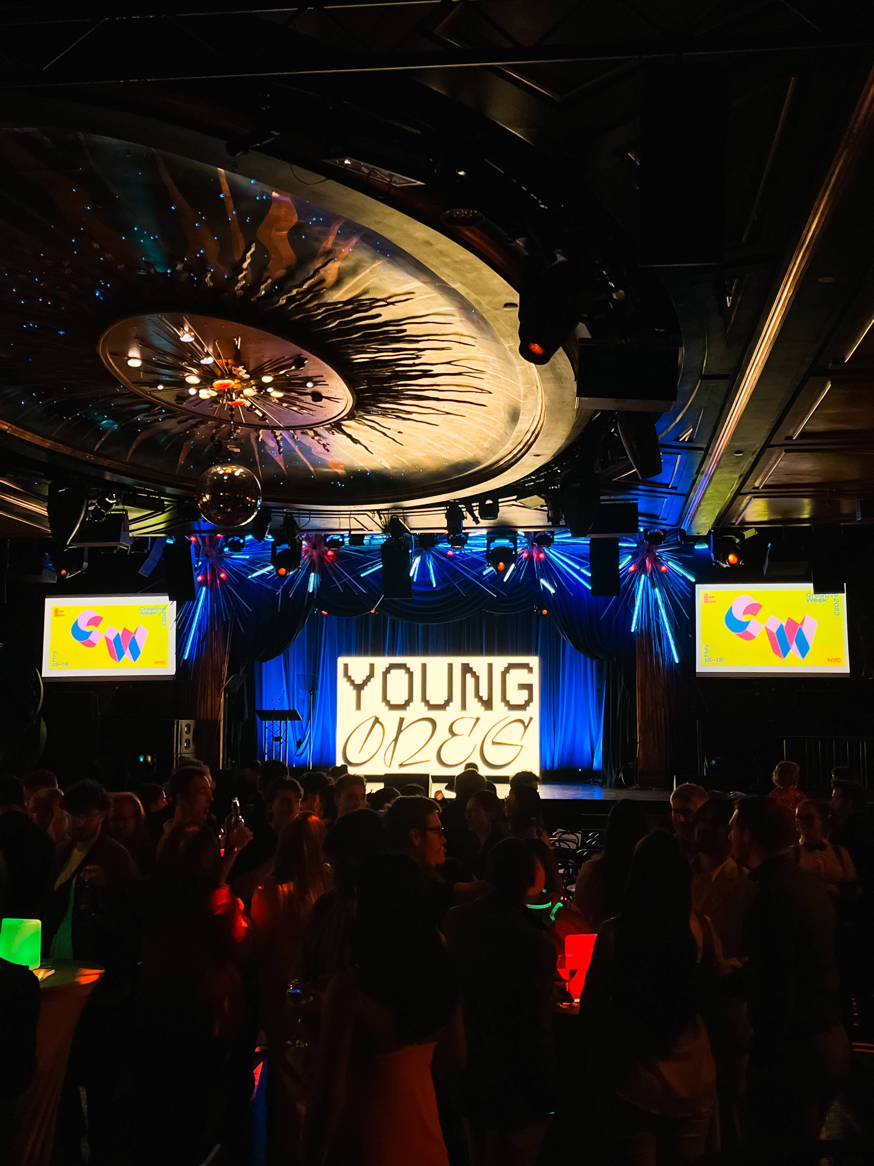 Un ambiente festivo con una multitud reunida frente a un escenario decorado con luces y un cartel que dice 'YOUNG'.