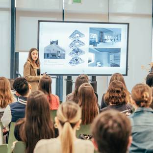 Una presentación educativa donde dos ponentes explican un proyecto arquitectónico frente a un público sentado.