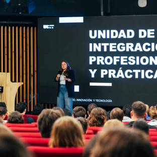 Una mujer presenta en un evento ante una audiencia sentada en un auditorio moderno.