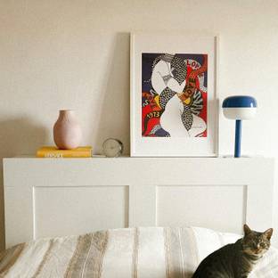 Una habitación con una cama, un gato sentado, una lámpara y una obra de arte en la pared.