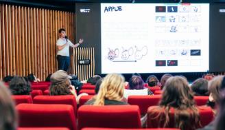 Una presentación en un auditorio con un ponente frente a un proyector que muestra ilustraciones.