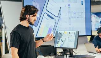 Un hombre presenta un diseño de interfaz de usuario frente a un monitor en una reunión de trabajo.