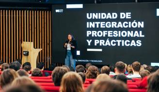 Una mujer presenta en un evento ante una audiencia sentada en un auditorio moderno.