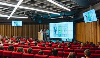 Una sala de conferencias donde se presenta un proyecto ante un público sentado en bancas rojas.