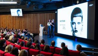 Una presentación en un auditorio con un orador y una proyección en pantalla que muestra una imagen de una modelo.