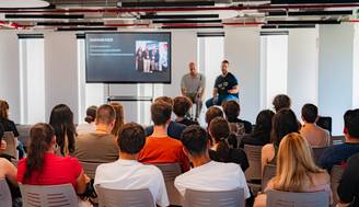 Un evento en una sala con un ponente y una audiencia sentada frente a una presentación en pantalla.