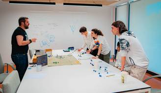 Un grupo de personas se reúne alrededor de una mesa mientras discuten y juegan con dados y figuras en un ambiente de trabajo colaborativo.