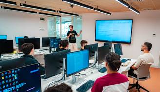 Un grupo de estudiantes escucha a un profesor en una sala de clases con computadoras.
