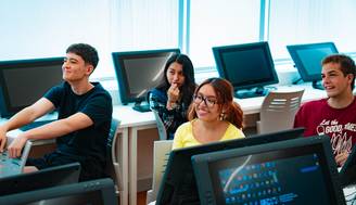 Un grupo de jóvenes sonríe mientras están sentados frente a computadores en un aula.