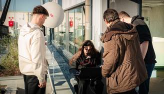 Un grupo de cuatro personas observan a una persona sentada frente a una computadora portátil en un entorno exterior.