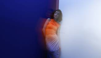 Una mujer posando con un vestido de tonos claros frente a un fondo de colores azul y blanco difuminados.