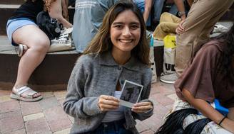 Una joven sonriente sostiene una foto mientras está sentada en un lugar con otras personas alrededor.