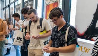 Un grupo de jóvenes se encuentra en un evento, mirando sus dispositivos móviles y sosteniendo bolsas de tela.