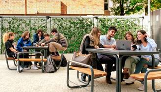 Un grupo de estudiantes se encuentra en un ambiente al aire libre, algunos trabajando en una laptop mientras otros conversan.