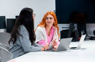 Dos jóvenes conversando en una mesa de trabajo con computadoras portátiles.
