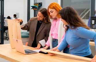 Tres mujeres observan una computadora portátil y discuten sobre un objeto de madera en un espacio de trabajo moderno.