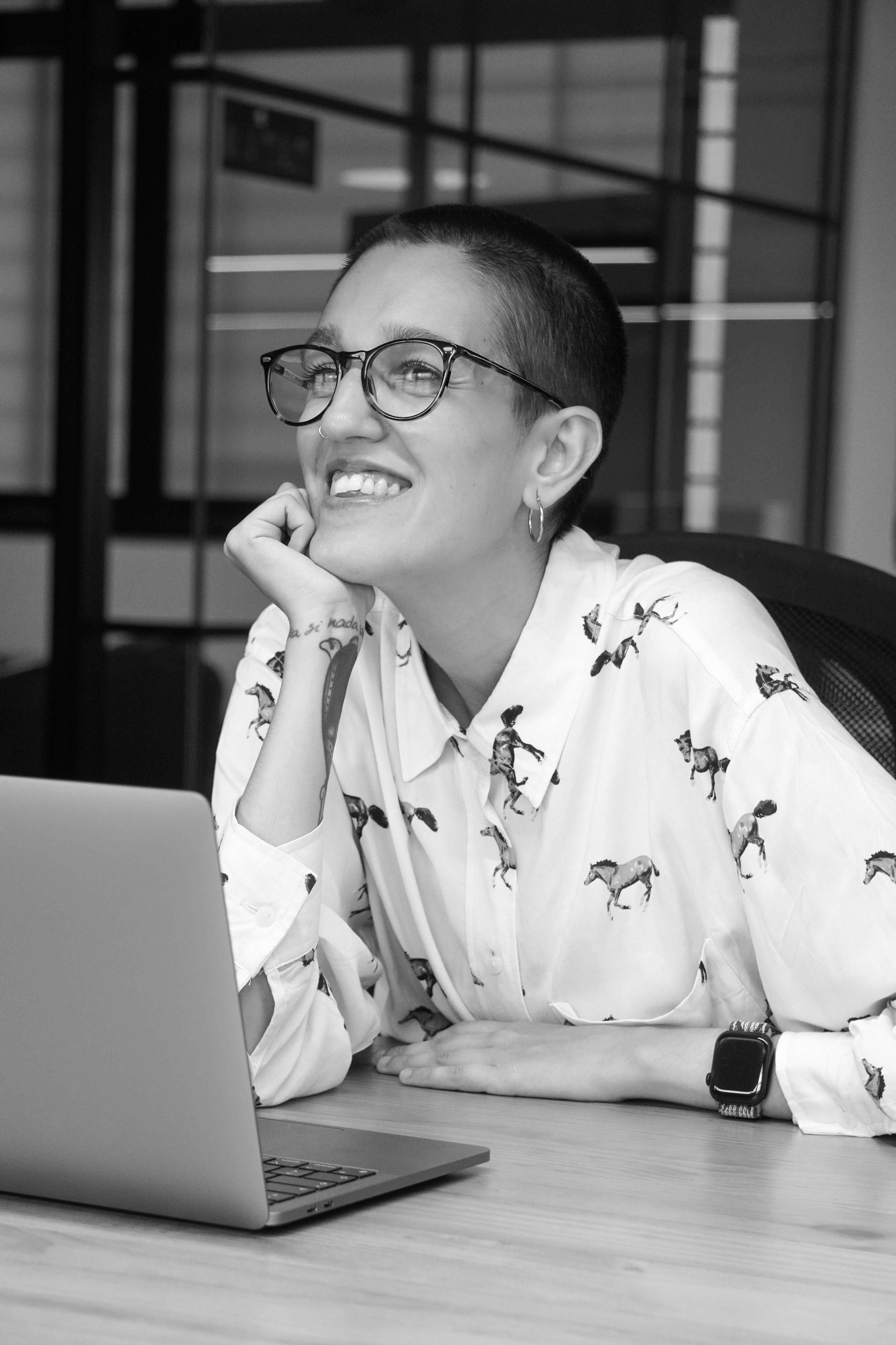 Una mujer sonriente con gafas se sienta frente a una computadora portátil en un ambiente de oficina.