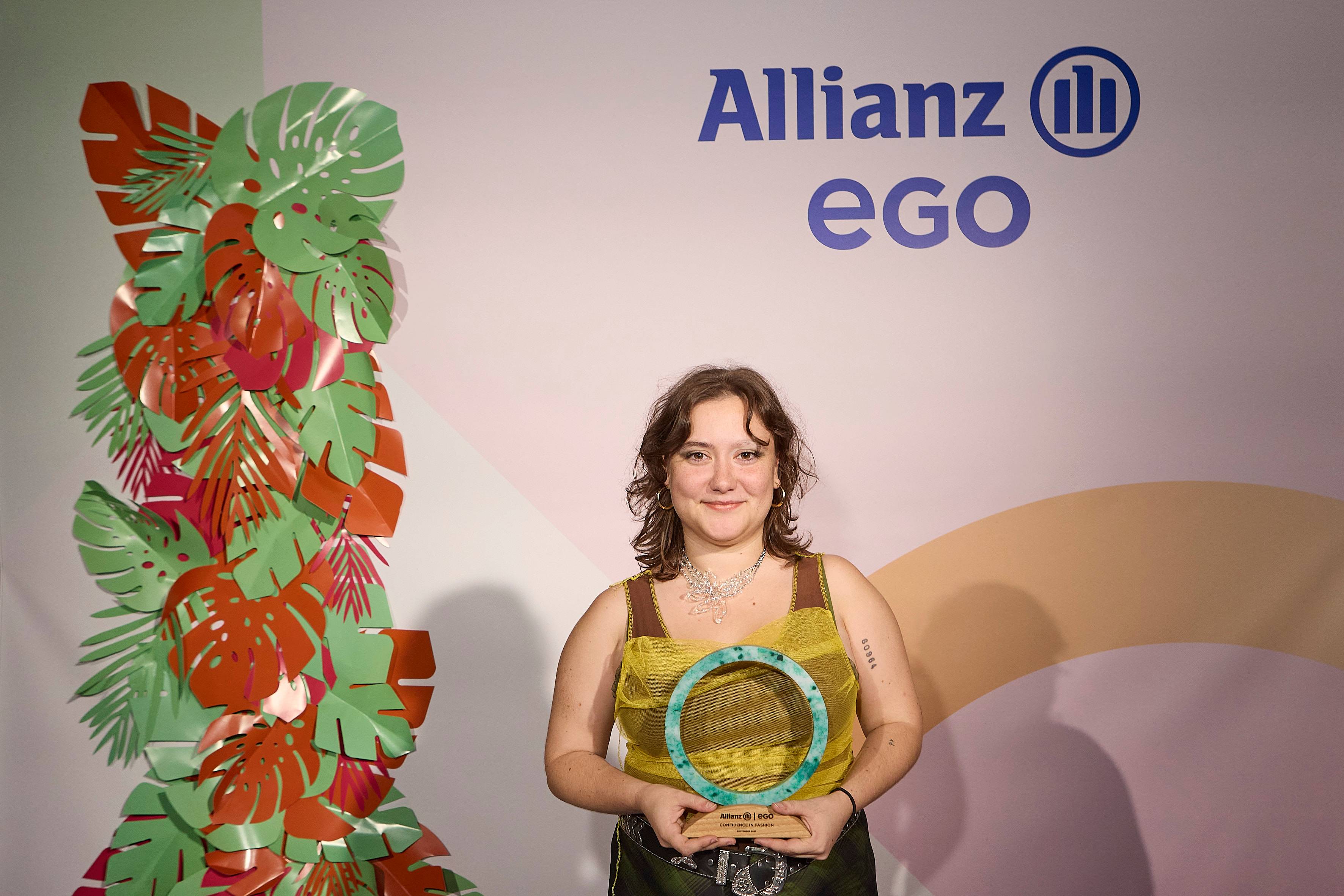 Una persona sonriente sostiene un trofeo frente a un fondo decorado con hojas de papel coloridas y el logotipo de Allianz EGO.