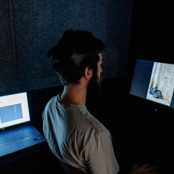 Un hombre sentado frente a dos pantallas de computadora en una habitación oscura, con un dispositivo en su cabeza.