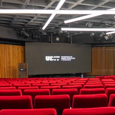 La imagen muestra un auditorio con filas de asientos rojos y una pantalla que exhibe el logo de la universidad.