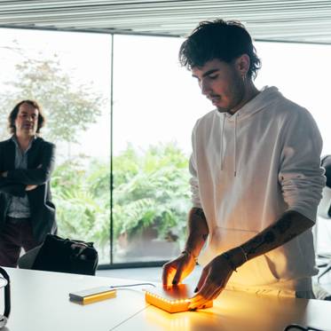 Un joven trabaja con un dispositivo luminoso en una mesa, mientras un hombre sonríe al fondo.
