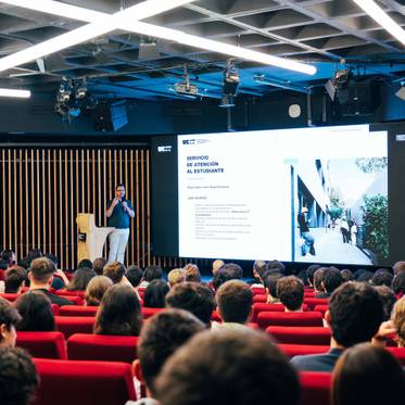 Un orador presenta en un auditorio a un grupo de estudiantes mientras se proyecta una diapositiva en una gran pantalla.