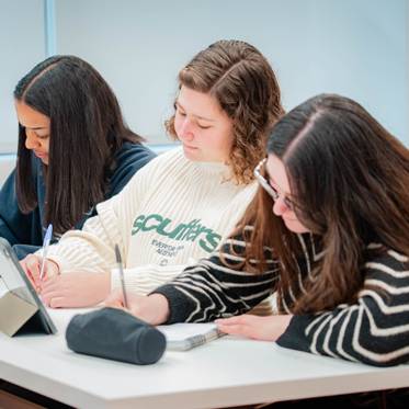Un grupo de tres estudiantes están concentrados tomando apuntes en un entorno de estudio moderno.