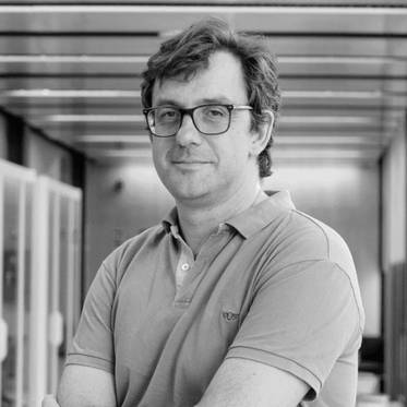 Retrato en blanco y negro de un hombre de pie con brazos cruzados, usando gafas y una camiseta de polo.