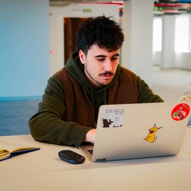 Un joven sentado frente a una computadora portátil en un entorno de oficina moderna.