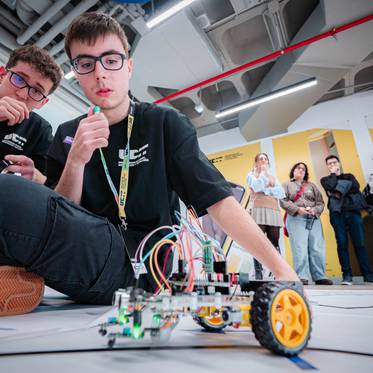 Dos jóvenes estudiantes están trabajando en un robot en una competición de robótica mientras otros observan al fondo.