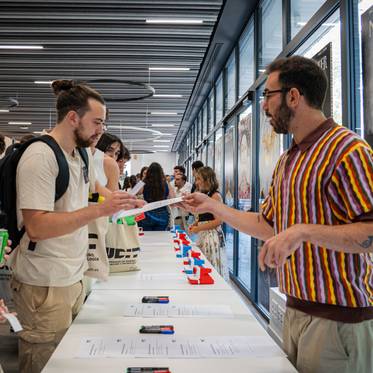 Un grupo de personas interactúa en un evento, mientras dos hombres conversan y exchangean documentos en un ambiente moderno y luminoso.