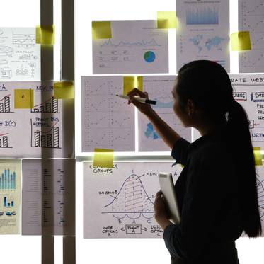 Una persona analiza gráficos y documentos en una presentación frente a una pared llena de notas y gráficos.