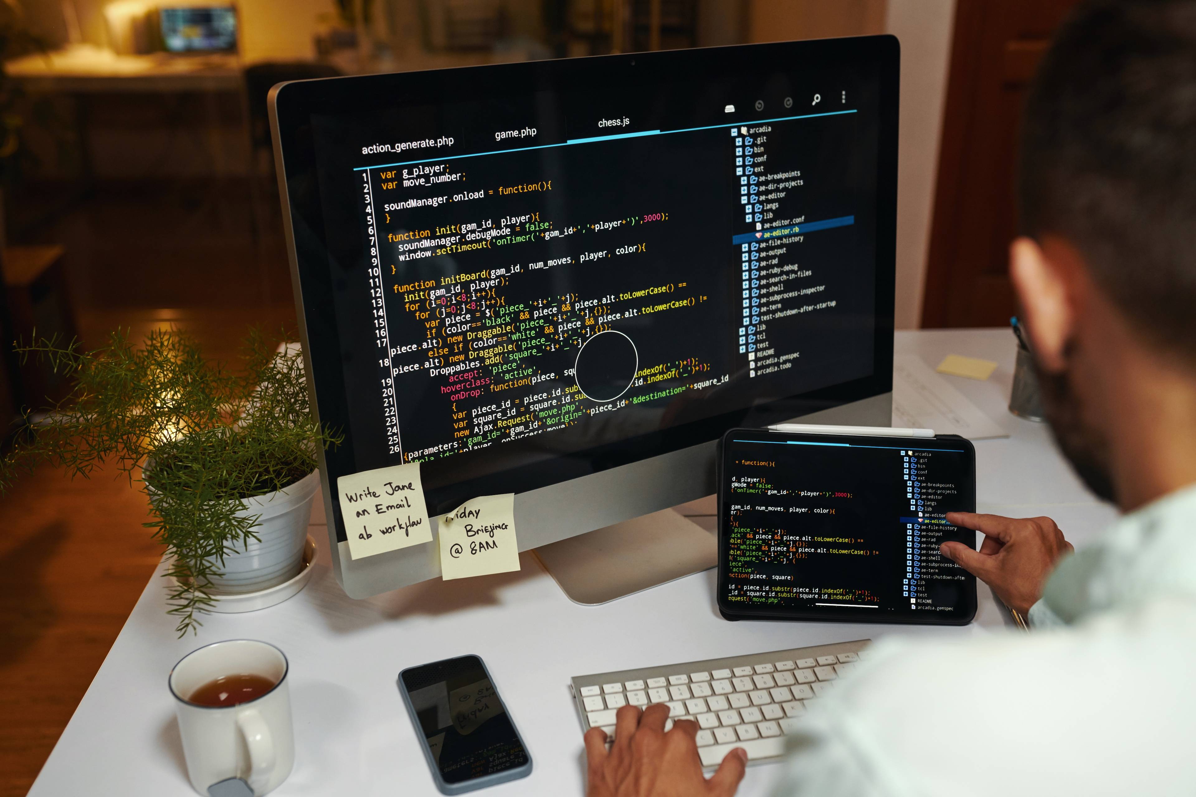 Una persona trabaja en un escritorio usando una computadora y una tablet, rodeada de café y plantas.