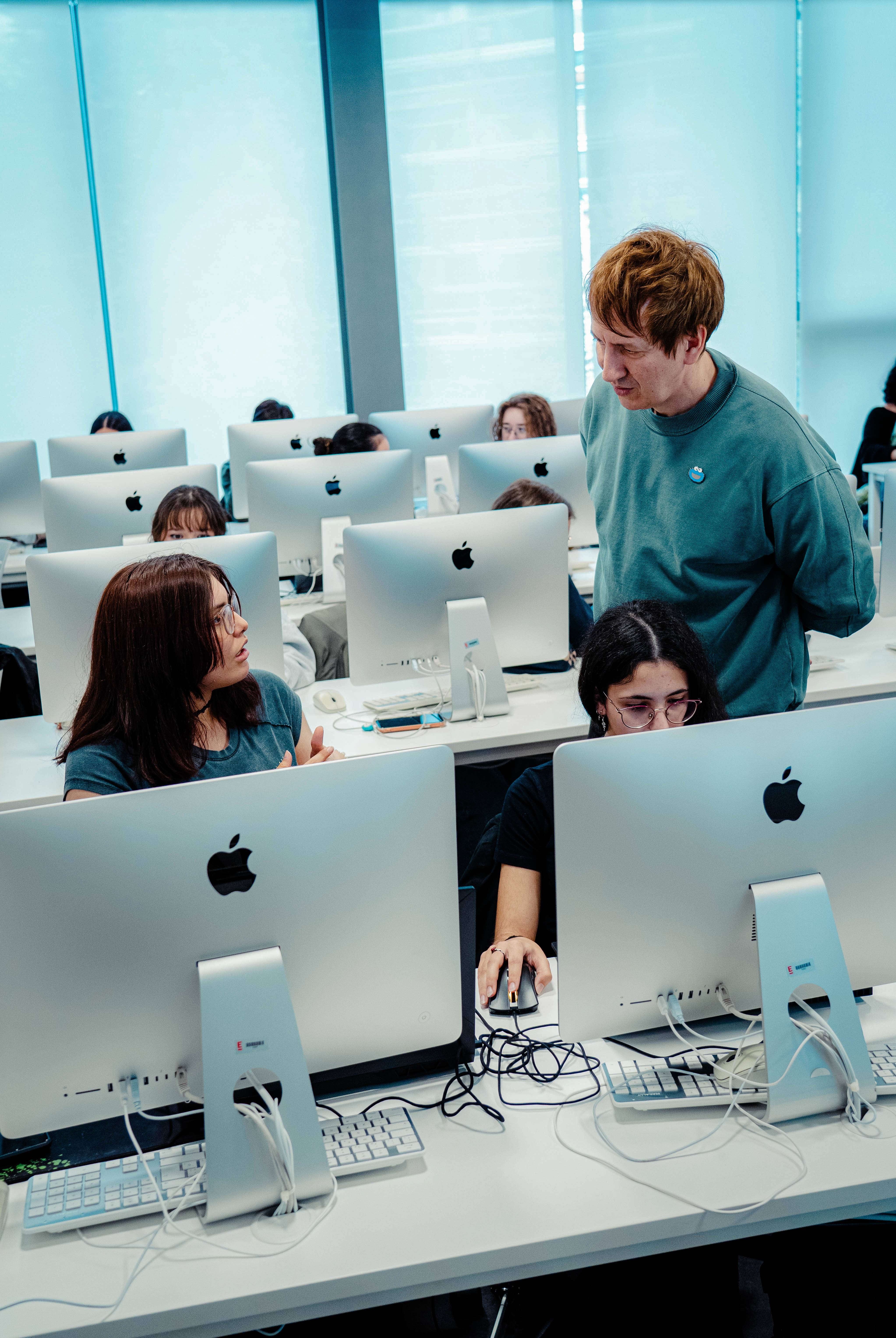 Grupo de estudiantes en un aula utilizando computadoras Apple mientras un instructor les supervisa.