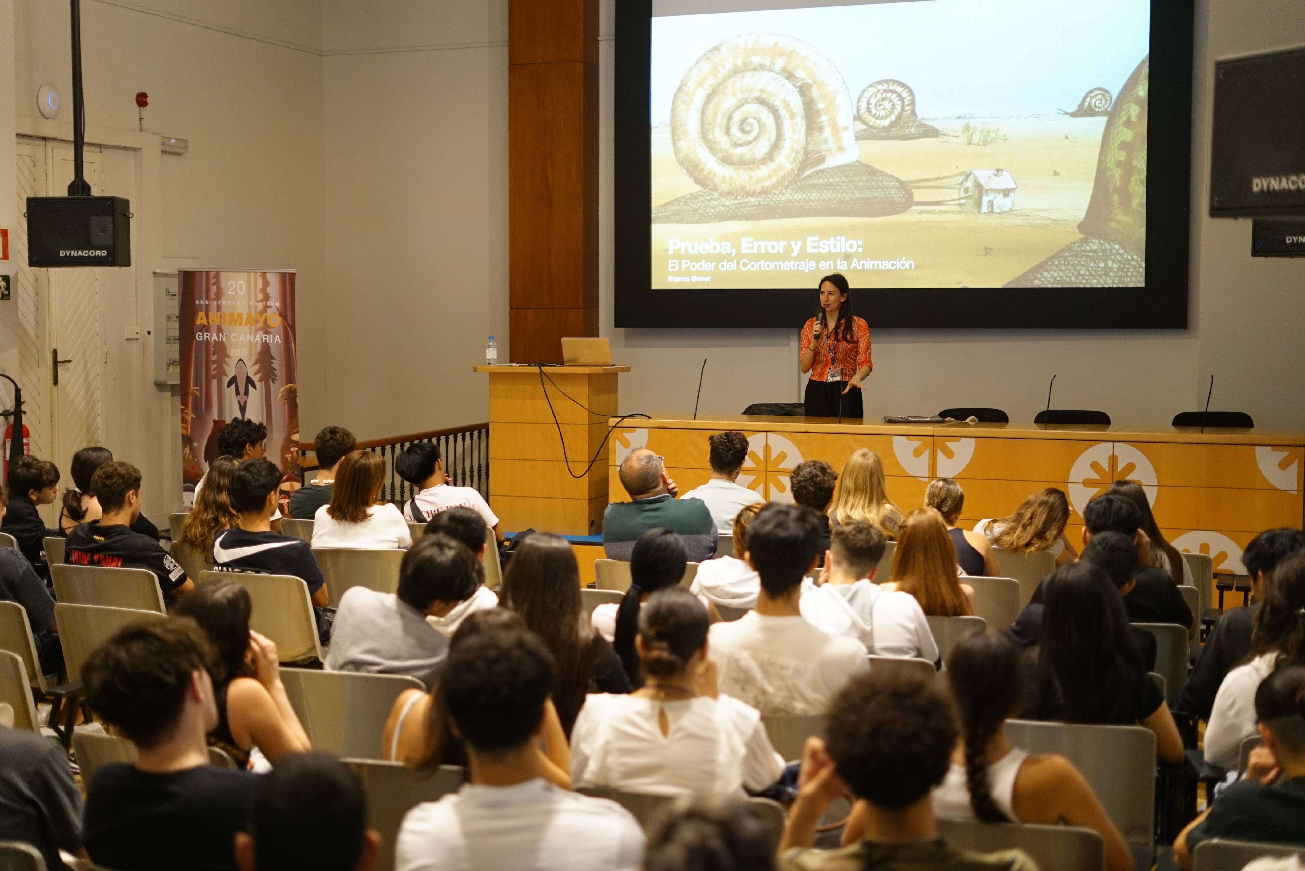 Una presentadora habla frente a una audiencia en una sala con proyección de imágenes en el fondo.