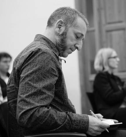 Un hombre con barba escribe en una libreta mientras otros lo observan en un ambiente de reunión.
