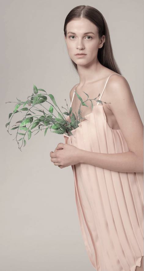 Una mujer con un vestido plisado de color rosa sostiene unas ramas verdes.