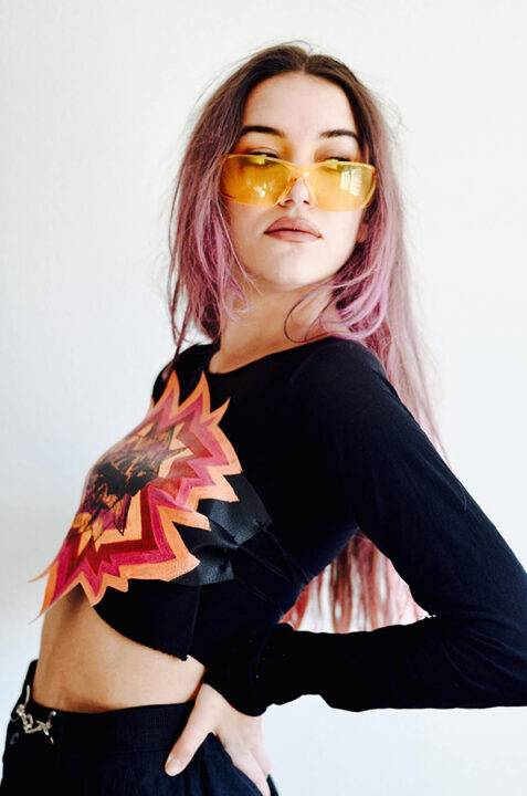 Una joven con cabello largo y rosado luce una camiseta negra con un diseño colorido y gafas de sol amarillas.