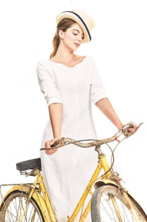 Una mujer con un vestido blanco y un sombrero posando junto a una bicicleta amarilla.