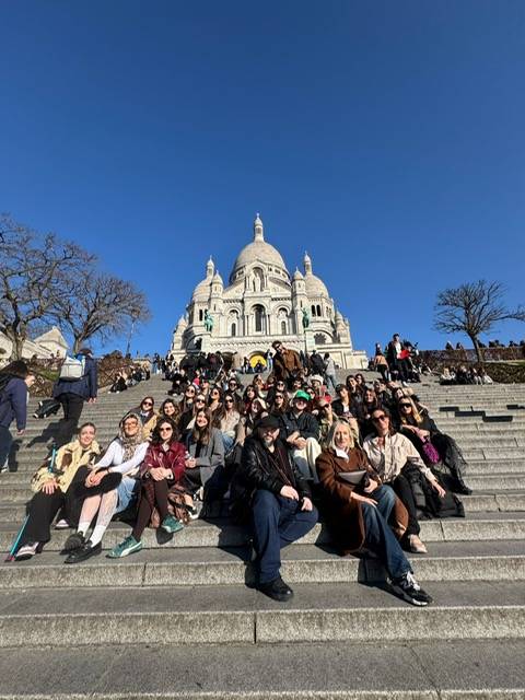 Un gran grupo de personas se sienta en las escaleras frente a la basílica del Sagrado Corazón en un día soleado.