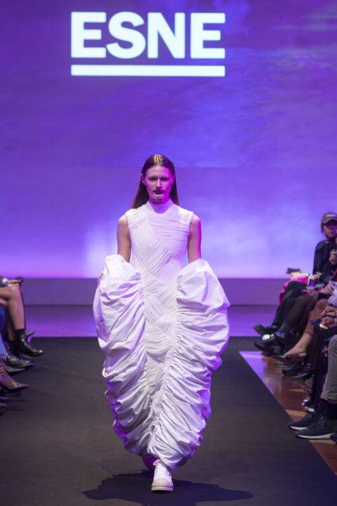 Una modelo presenta un vestido blanco con un diseño voluminoso en una pasarela iluminada con luces moradas.