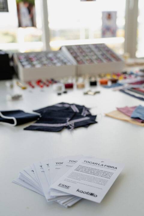 Una mesa con muestras de tejidos y una caja con botones, acompañada de folletos informativos sobre el evento 'Tocar la fibra'.