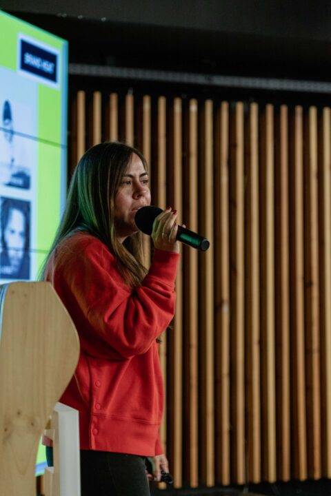 Una mujer habla en un micrófono en un escenario con un fondo de panel de madera.
